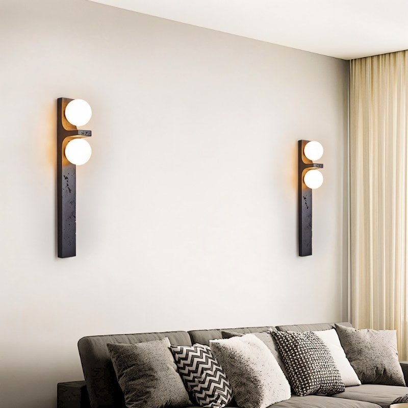 Two Clérachic black wall lamps with frosted globes hung on a pale living room wall above a grey sofa and cushions.
