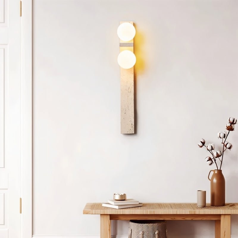 Clérachic travertine wall lamp with two lit globes above a wooden console table with cotton stems and a ceramic vase.
