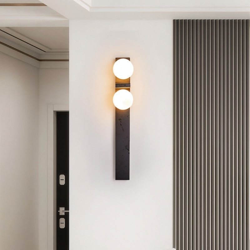 Clérachic black wall lamp with two warm glass globes in a hallway, beside a ribbed grey wall panel and a dark door.
