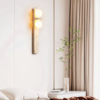 Clérachic travertine wall lamp glowing beside a bed with sheer curtains, a slim vase of green stems and stacked books.
