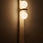 Close view of a Clérachic travertine wall lamp lit in a dim room, its pitted cream stone and two soft globes glowing.