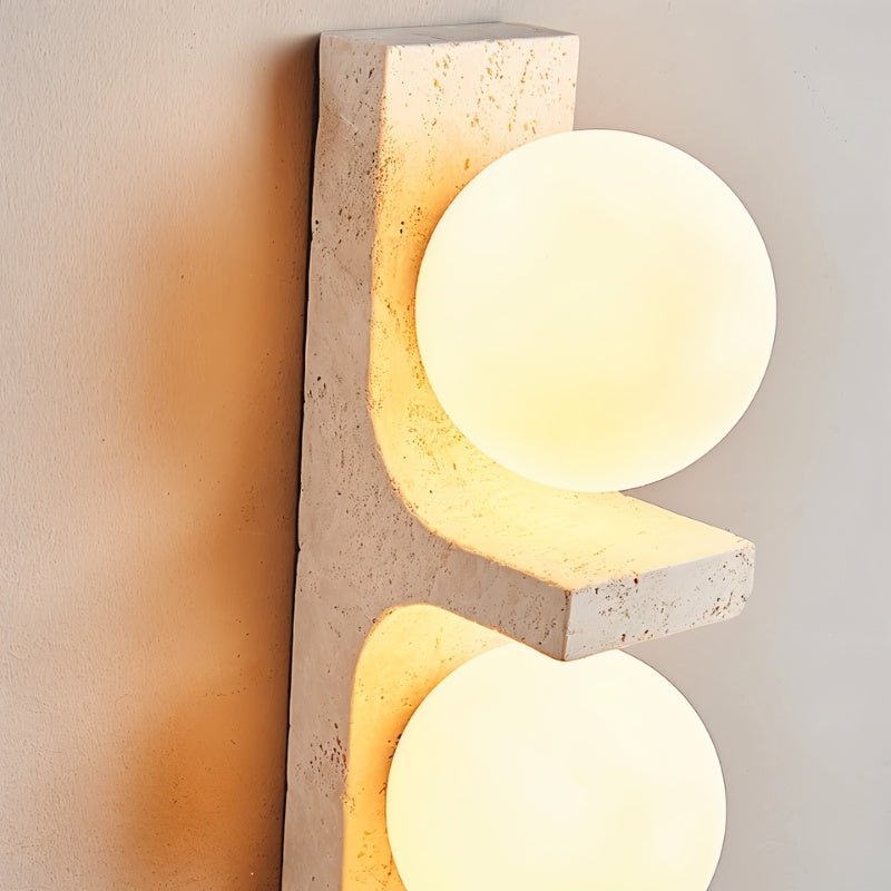 Close-up of the Clérachic travertine wall lamp: a pitted cream stone shelf holds two frosted white globes that glow warmly.