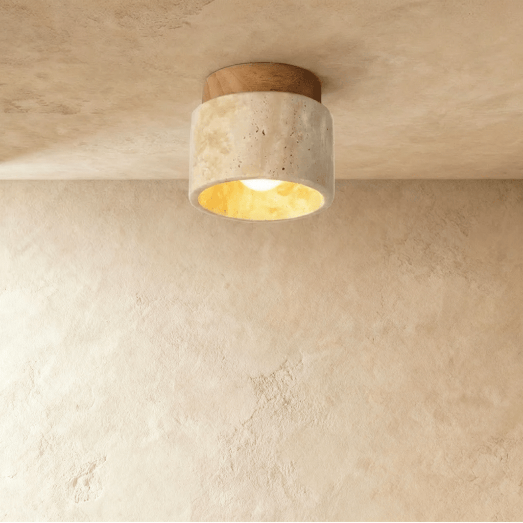 Clérachic short travertine cylinder ceiling light with a light wood canopy, bulb glowing warm on a limewashed plaster ceiling.