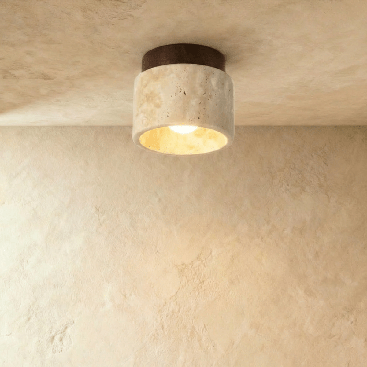 Clérachic short travertine ceiling light with a dark walnut canopy, the bulb lit warm inside the stone cylinder.