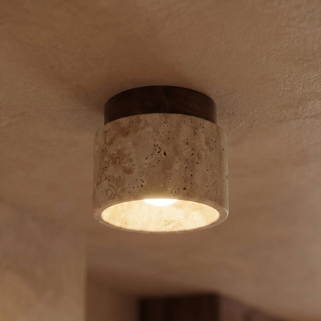 Close view of a Clérachic short travertine ceiling light, walnut canopy above porous stone, bulb glowing inside.