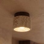 Close view of a Clérachic short travertine ceiling light, walnut canopy above porous stone, bulb glowing inside.