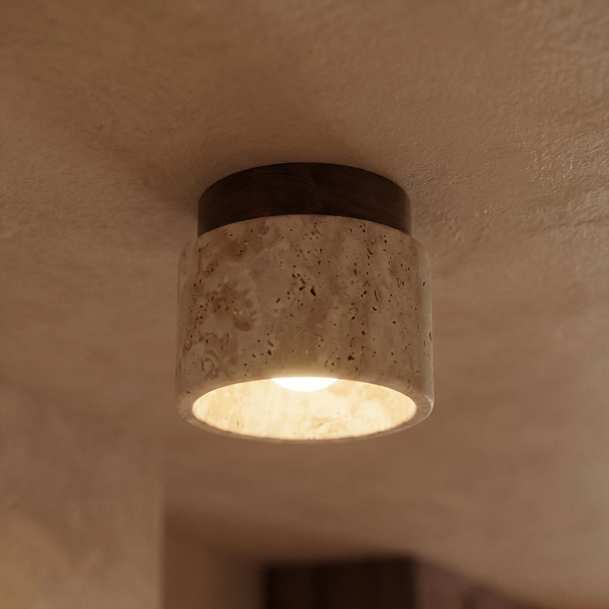 Close view of a Clérachic short travertine ceiling light, walnut canopy above porous stone, bulb glowing inside.