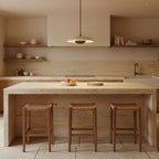 A single Clérachic travertine pendant hangs over a travertine kitchen island with three woven-seat wooden stools.