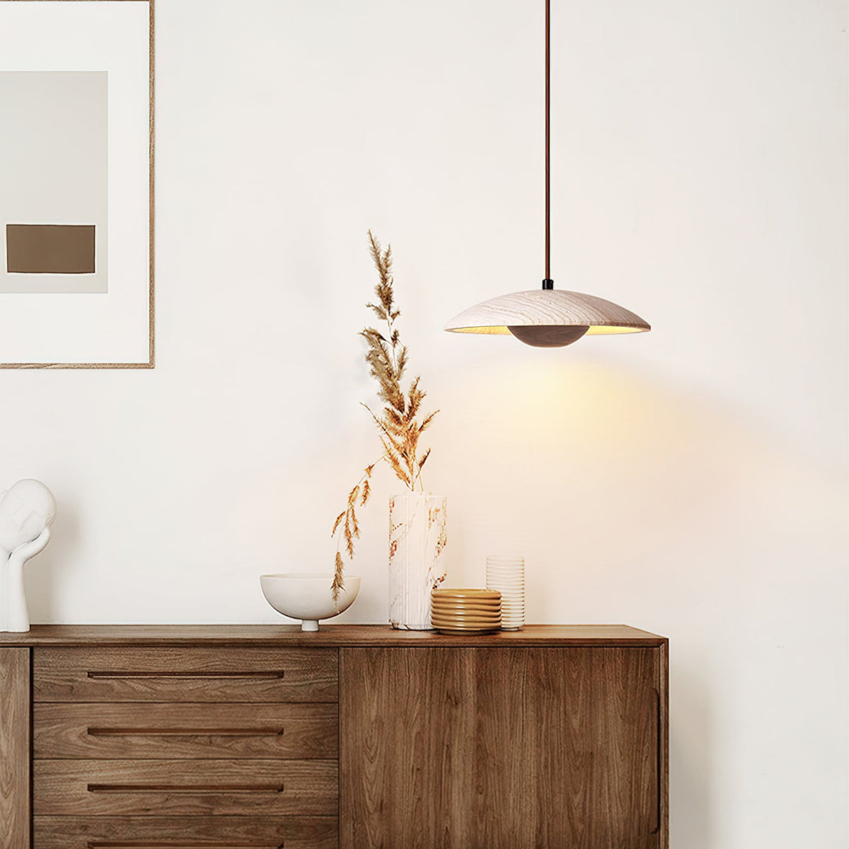 A Clérachic pendant with a travertine shade hangs beside a walnut sideboard with dried stems and a framed print.