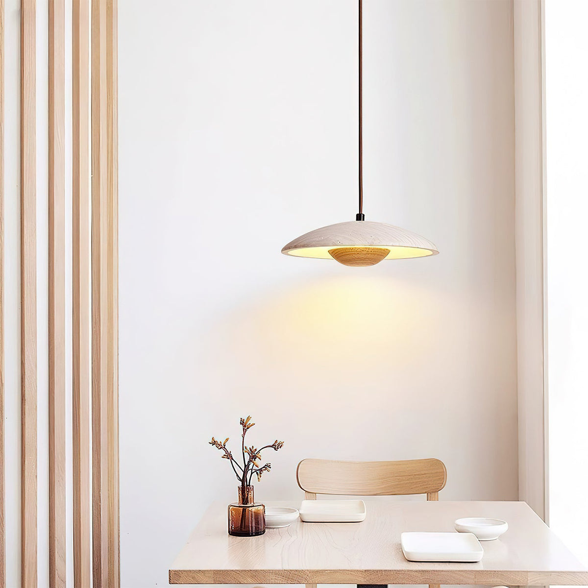 Clérachic travertine pendant hangs above a pale wood table with a sprig in a vase, beside slatted wood panelling.