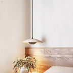 A Clérachic travertine pendant hangs beside a light wood headboard, with a vase of eucalyptus lit from below.