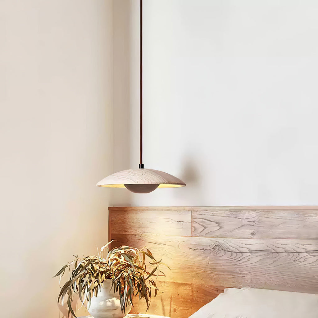 A Clérachic travertine pendant hangs over a headboard of pale wood, with a vase of dried leaves beneath it.