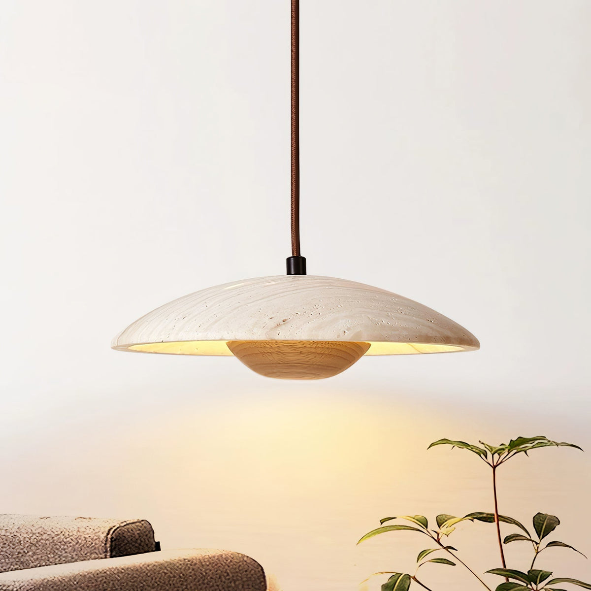 This Clérachic travertine pendant sits over a white wall with a houseplant and sofa edge, in warm light.