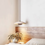 A Clérachic travertine pendant hangs by a bed with a wooden headboard, a vase of leaves in the warm light.