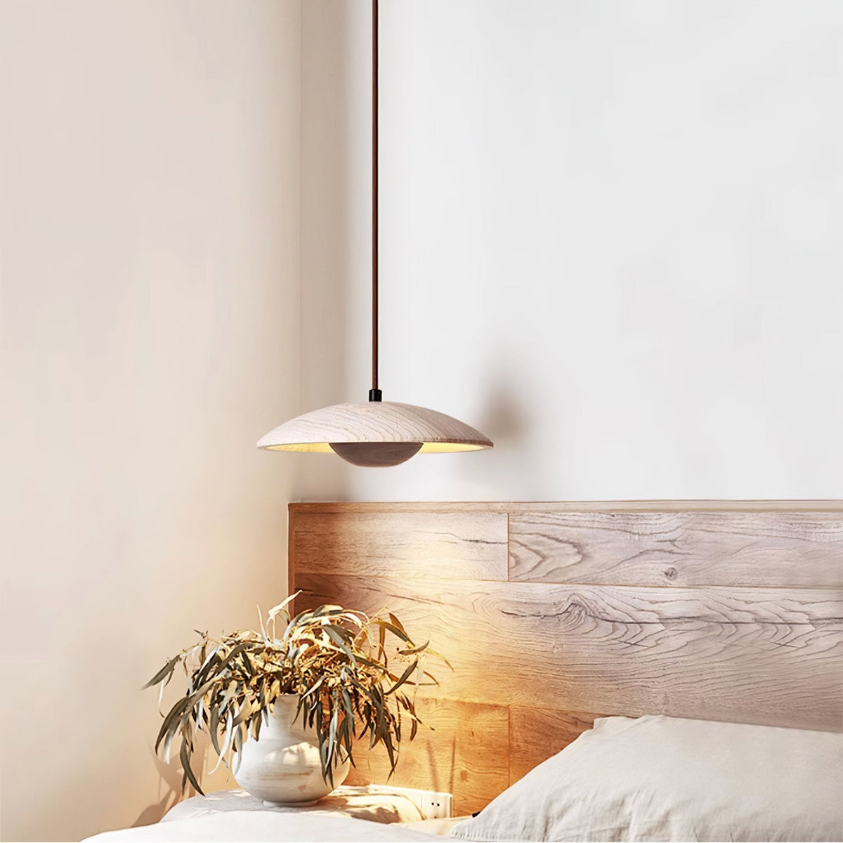 A Clérachic travertine pendant hangs by a bed with a wooden headboard, a vase of leaves in the warm light.