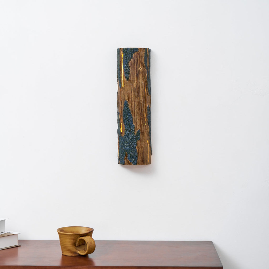 Hung high on a white wall, this Clérachic Bark wall light glows through its narrow slits, with a wooden cup on the sideboard below.