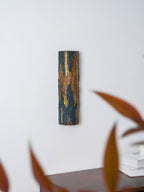 Behind soft russet leaves, a Clérachic Bark wall light with a lit slit stands out against a white wall above a wooden sideboard.