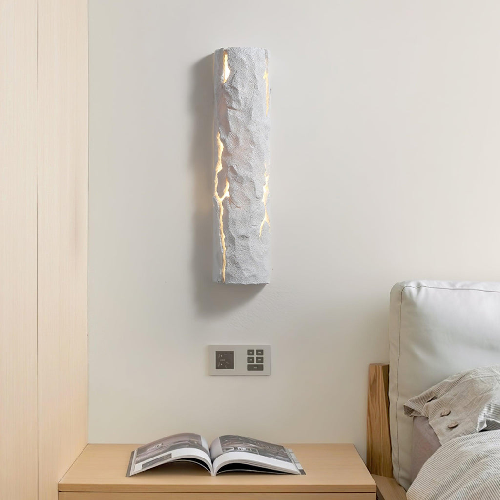 A tall white Clérachic wall light with a rough, chiselled surface glows through its cracks above a pale bedside shelf and an open book.