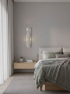 Seen across a calm bedroom, a white Clérachic wall light glows through its fine cracks above a pale wood bedside table.