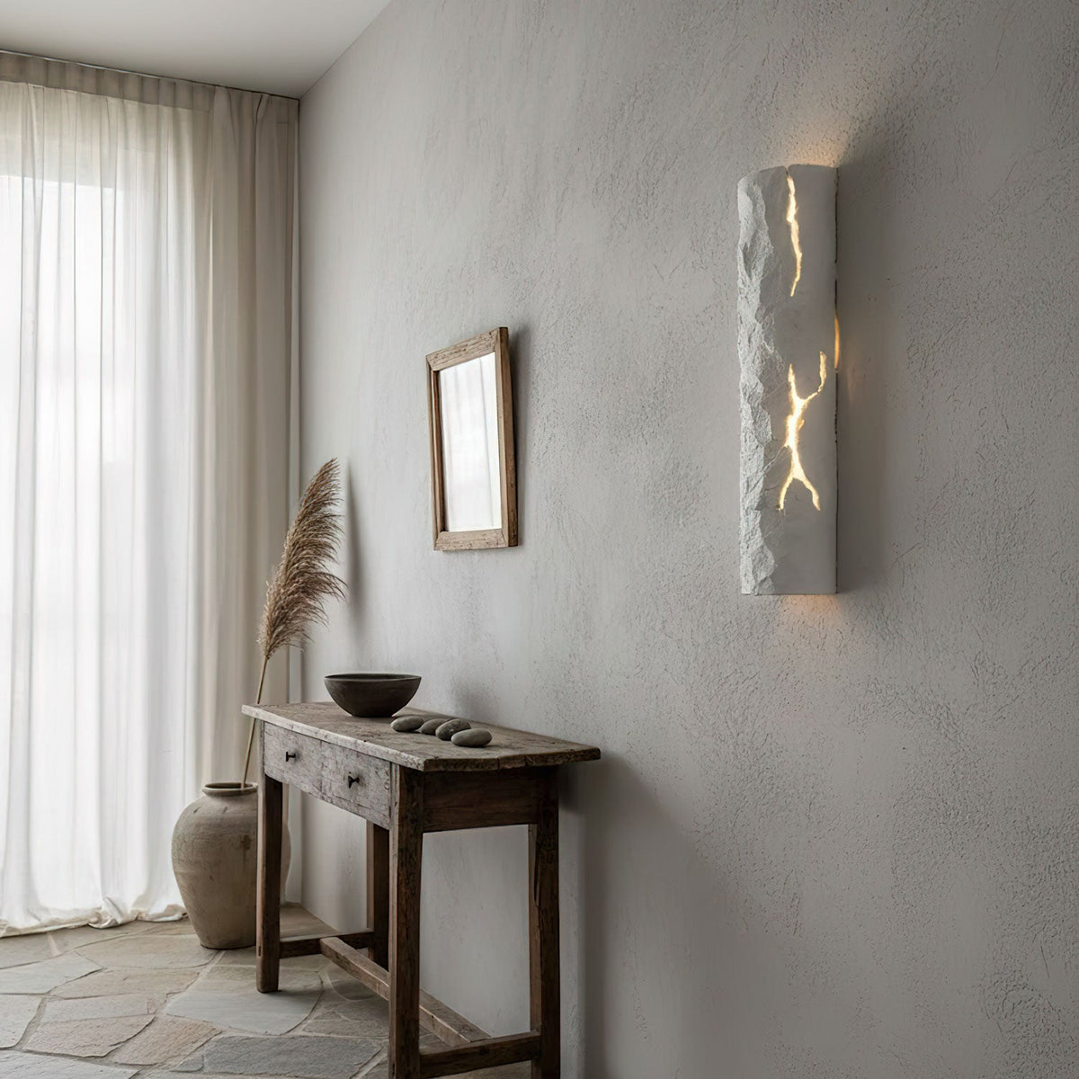 A white Clérachic wall light with a rough plaster-like surface lights up its cracks on a limewashed wall above a rustic console table.