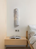 A narrow white Clérachic wall light, lit through its cracks, hangs above a wall switch and a pale bedside shelf with an open book.