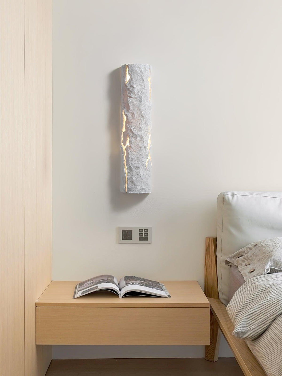 A narrow white Clérachic wall light, lit through its cracks, hangs above a wall switch and a pale bedside shelf with an open book.