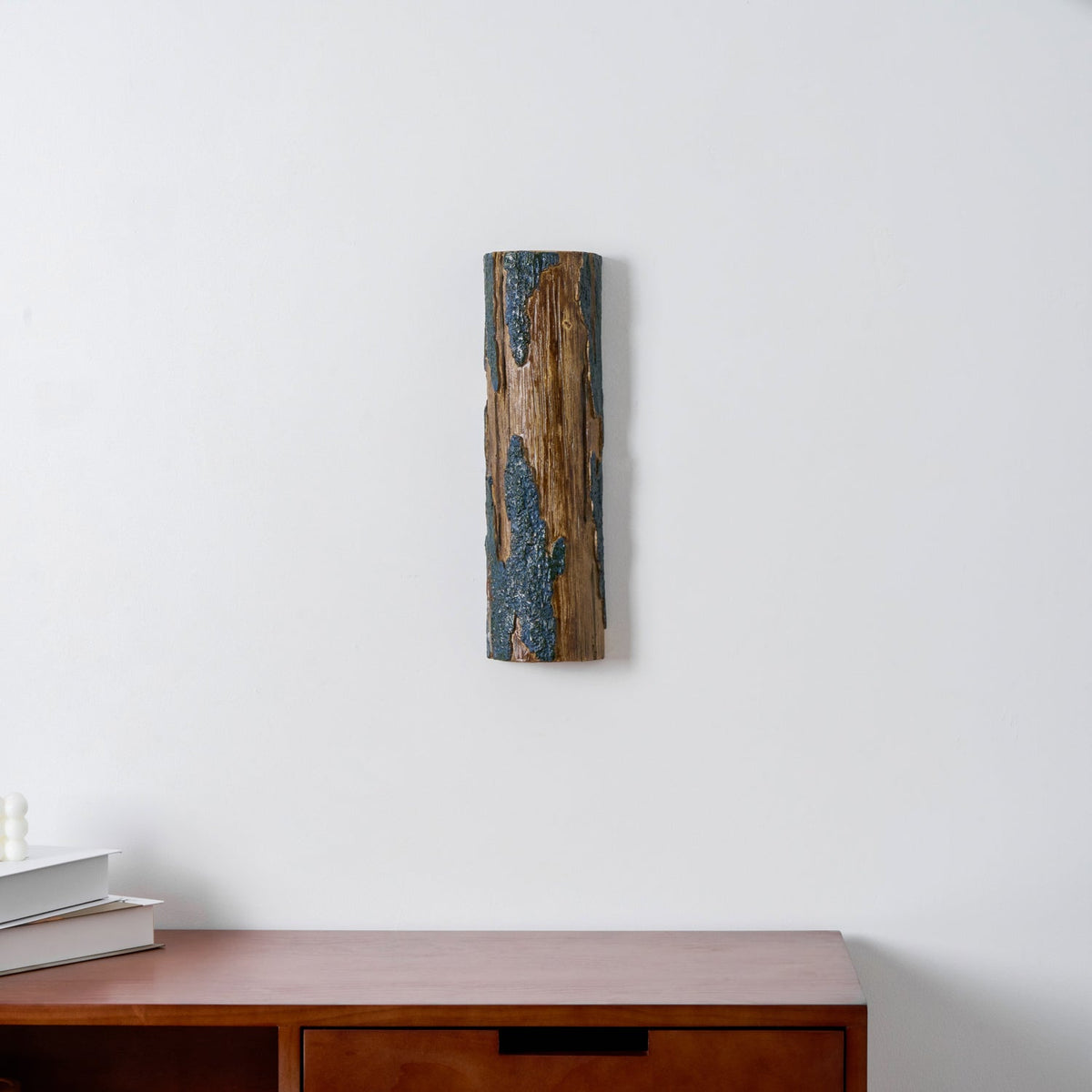 A Clérachic Bark wall light, unlit, hangs on a white wall above a wooden sideboard with a stack of books to one side.