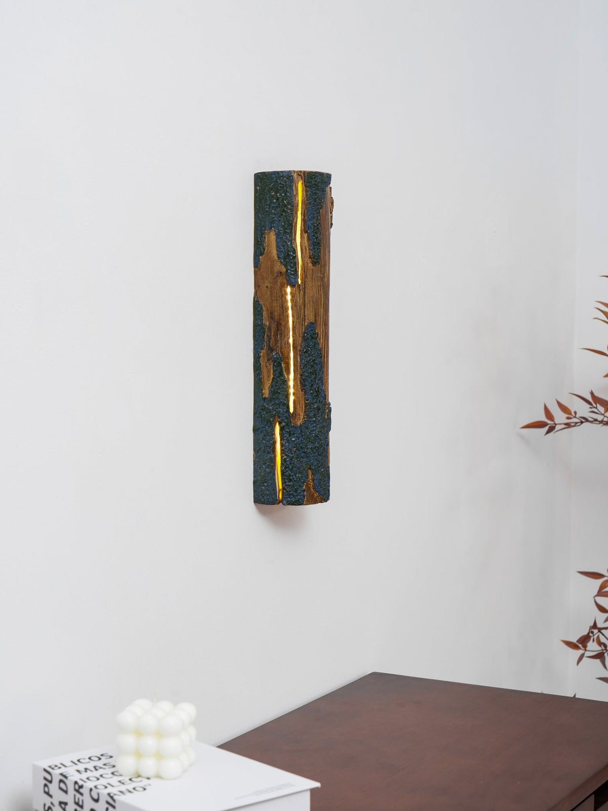 From a slight angle, a Clérachic Bark wall light with a glowing slit hangs on a white wall above a white cube candle and a wooden table.
