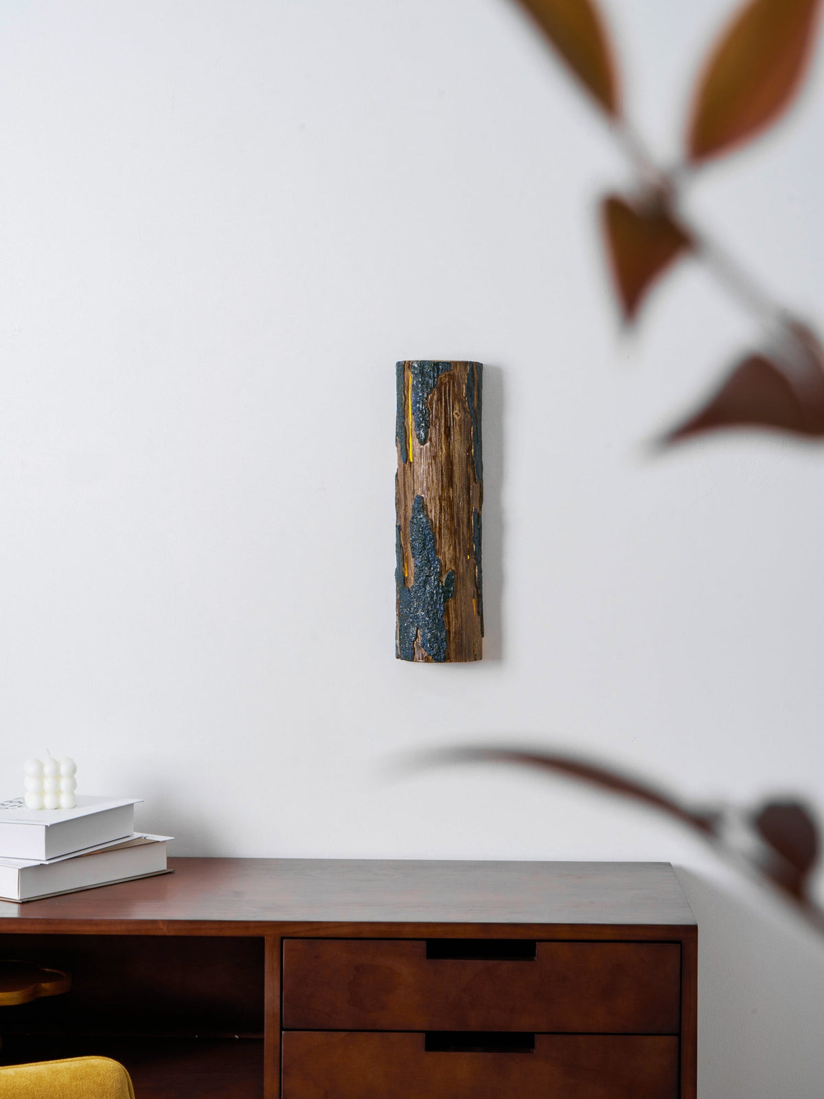 Through blurred russet leaves, a small Clérachic Bark wall light is visible on a white wall above a drawer unit and books.