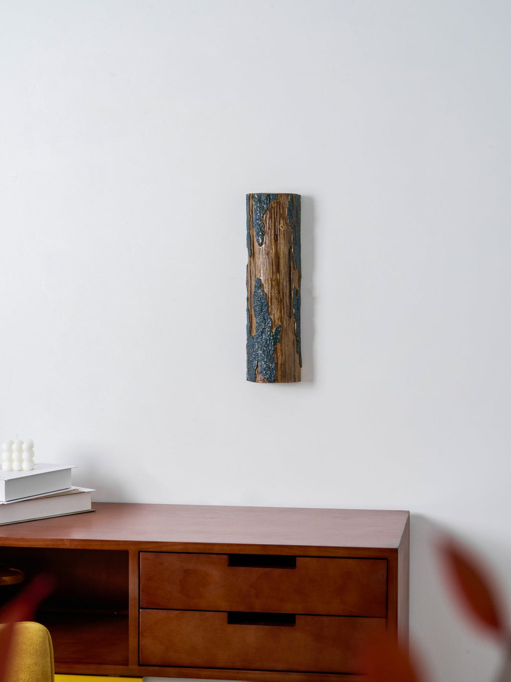 A Clérachic Bark wall light seen across a wooden drawer unit, with out-of-focus leaves in the foreground and a white wall behind.