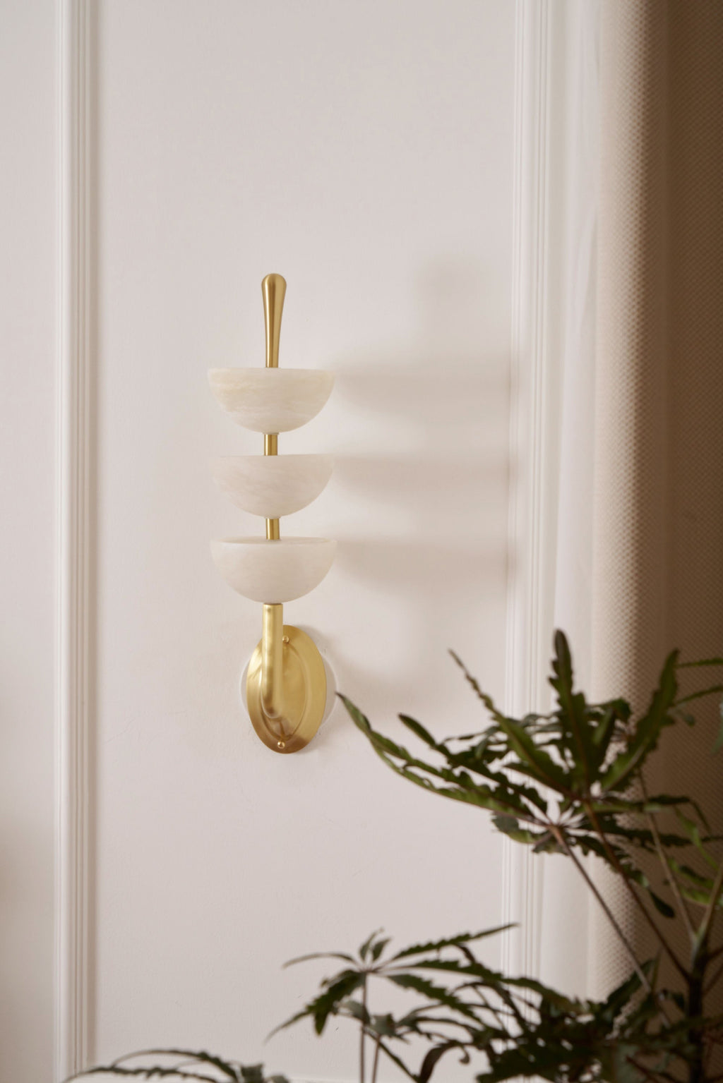 Clérachic triple-dome wall sconce mounted on a panelled cream wall beside a fern, brass bracket set on its oval backplate.