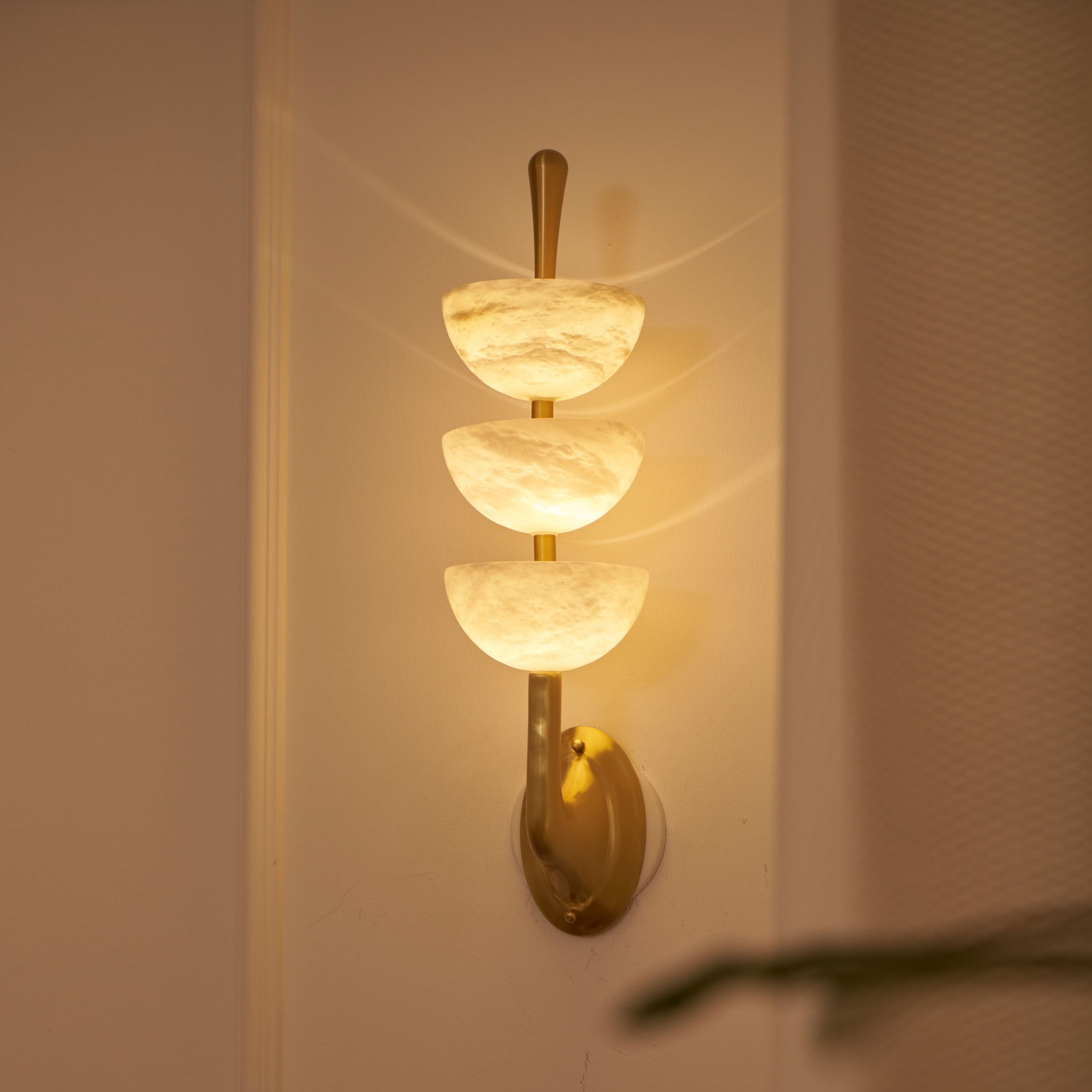 Clérachic triple-dome wall sconce lit at night, the alabaster turned honey-amber with soft arcs thrown across the panelling.