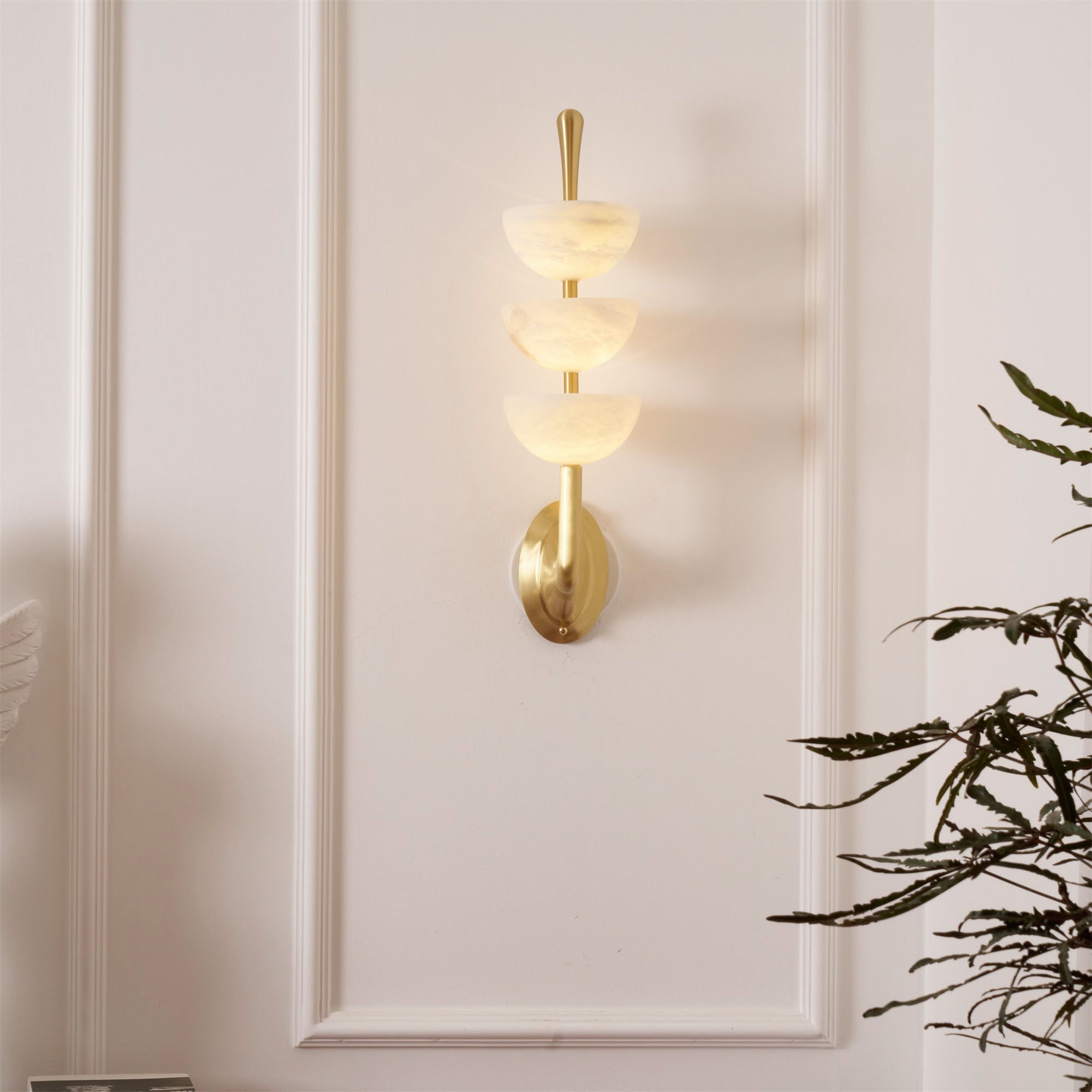 Clérachic triple-dome sconce glowing softly on a pale panelled wall, the three bowls lighting the run above and below.