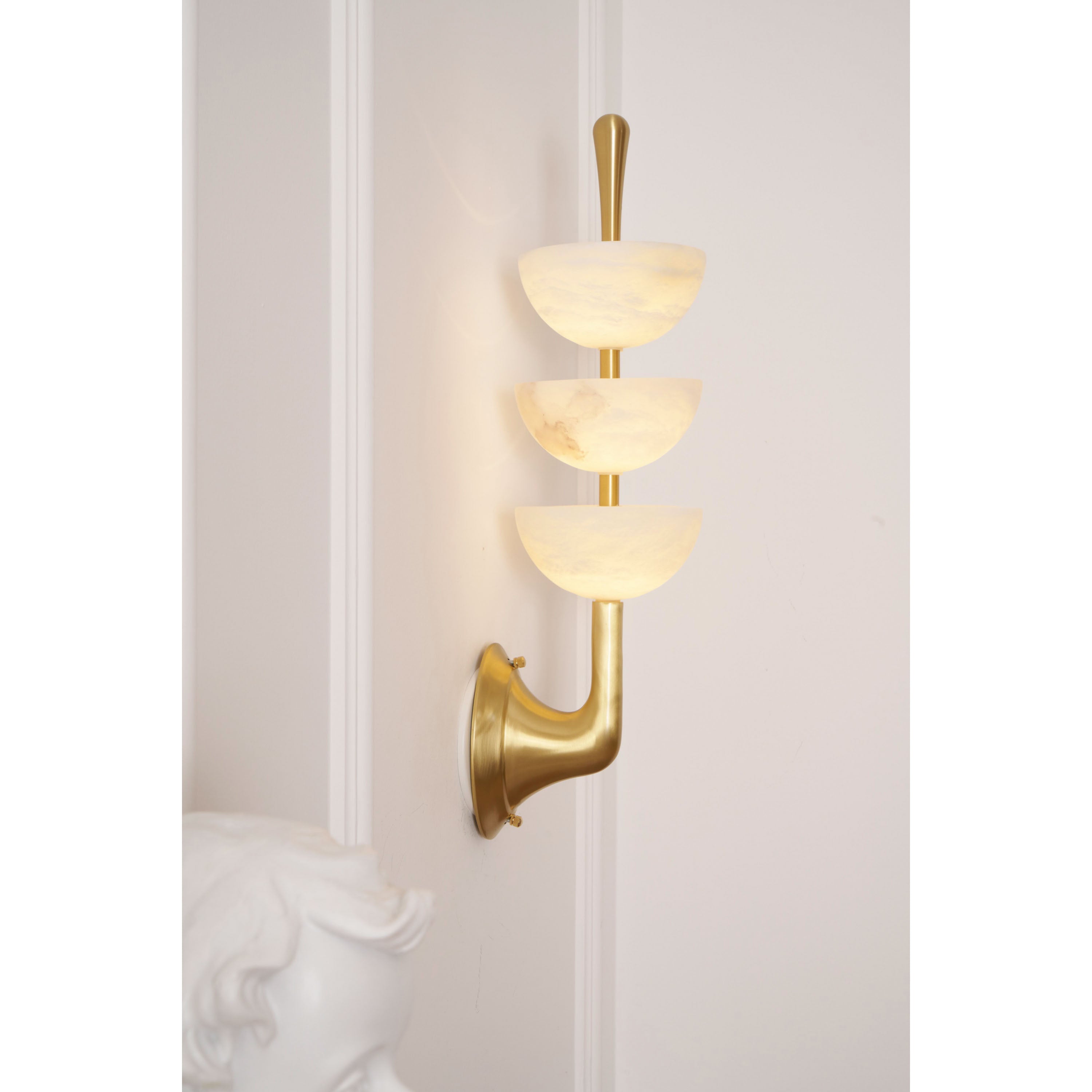 Angled view of the lit Clérachic wall sconce beside a plaster bust, brass arm and backplate catching the light on the wall.
