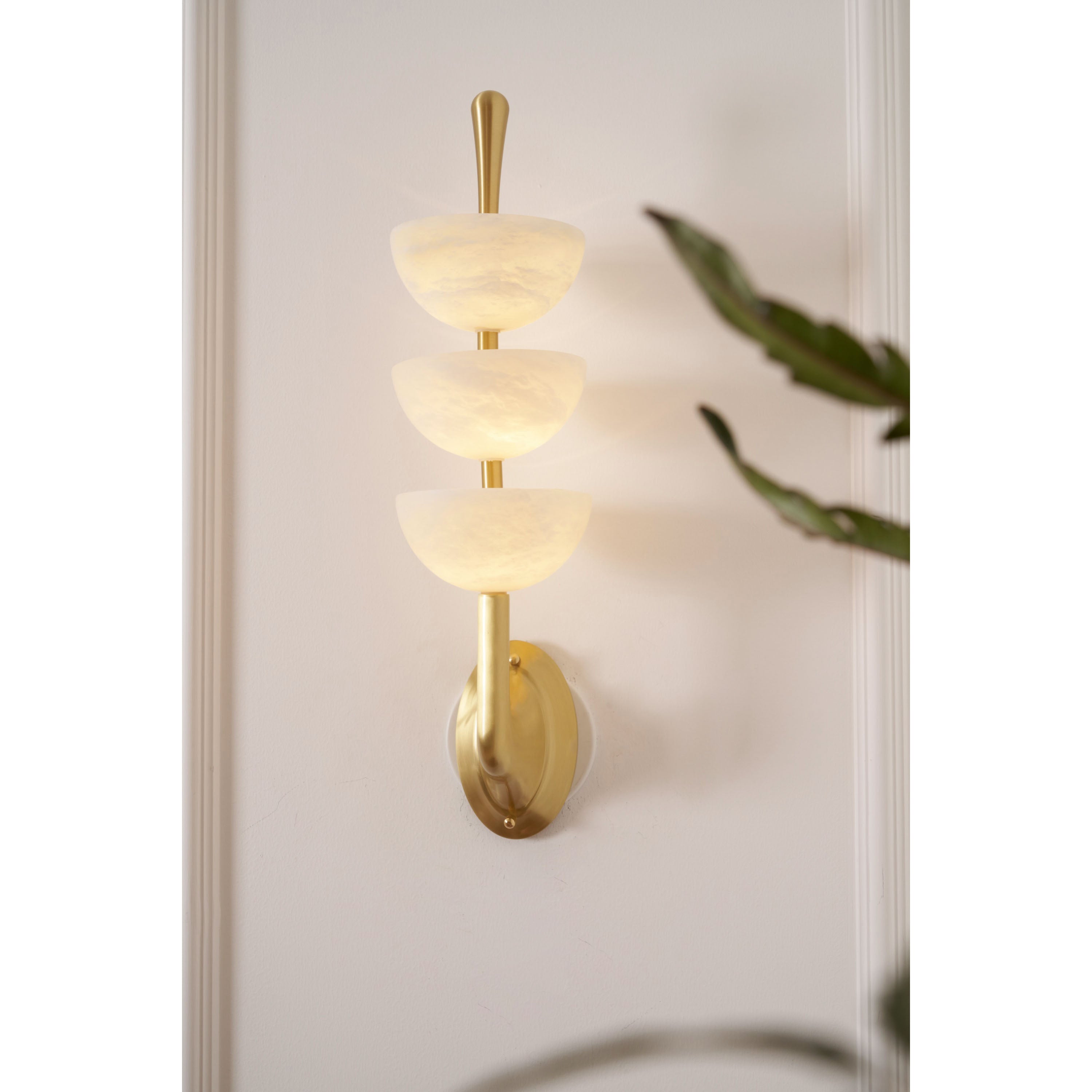 Clérachic triple-dome wall sconce lit beside a trailing houseplant, warm glow held inside the three stacked alabaster bowls.