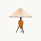 This Clérachic table lamp on a plain white backdrop, a pleated beige shade over a turned wood base and black tripod legs.