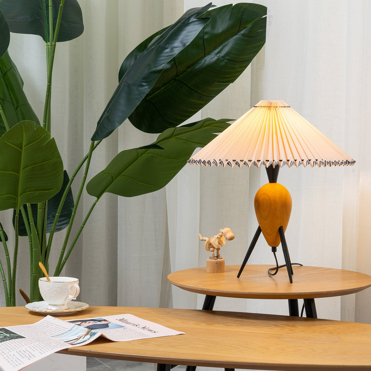 Beside a leafy plant and sheer curtains, a Clérachic table lamp stands on a round wooden side table with a toy dinosaur.