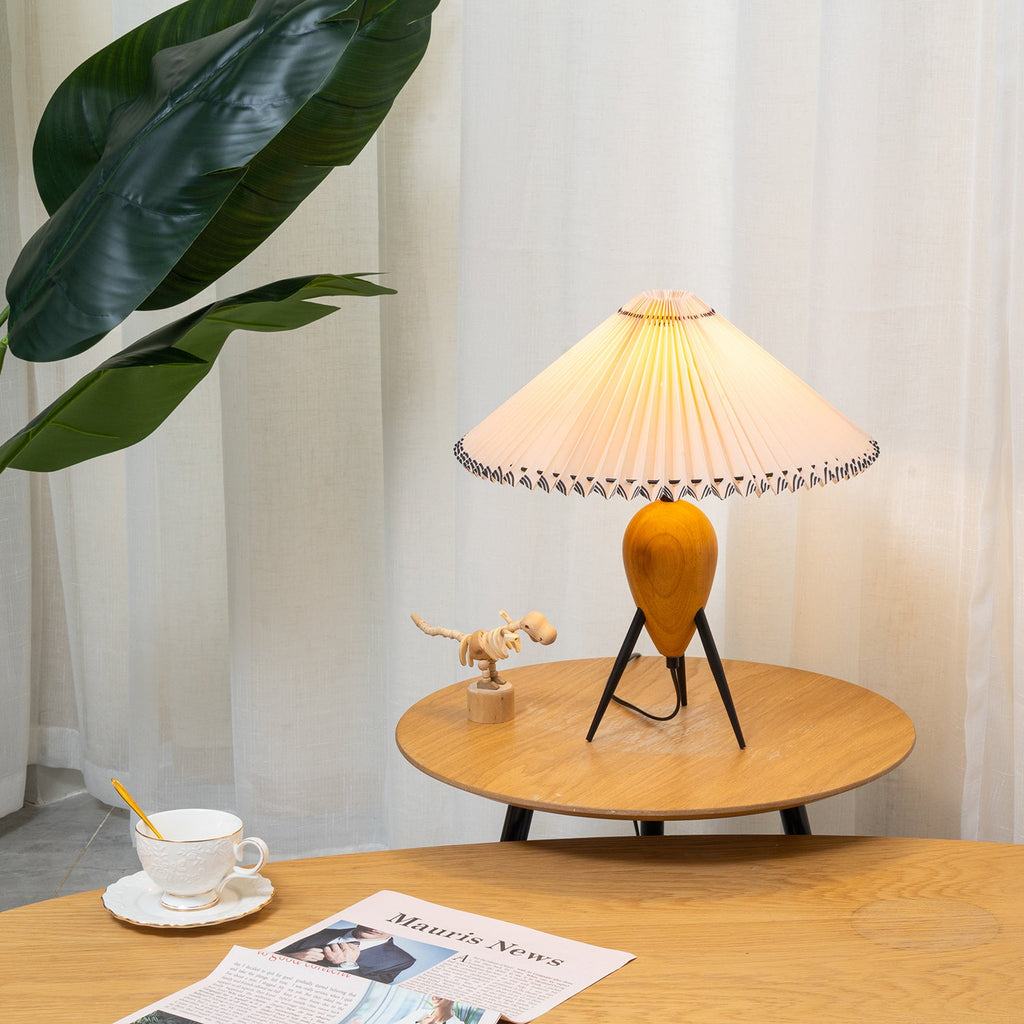 A Clérachic table lamp on a small round oak table, sheer curtains and a green plant behind, a cup and newspaper in front.