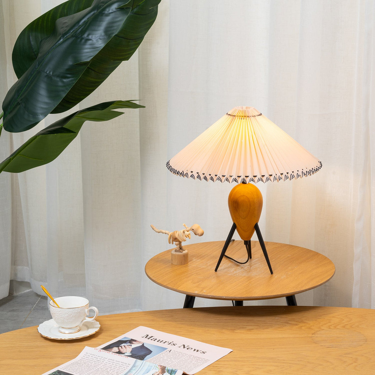 A Clérachic table lamp on a small round oak table, sheer curtains and a green plant behind, a cup and newspaper in front.