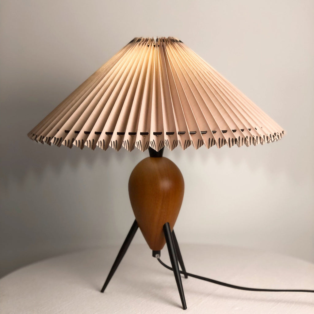 Close view of the wood base and black tripod legs of this Clérachic table lamp, under its wide pleated beige shade.