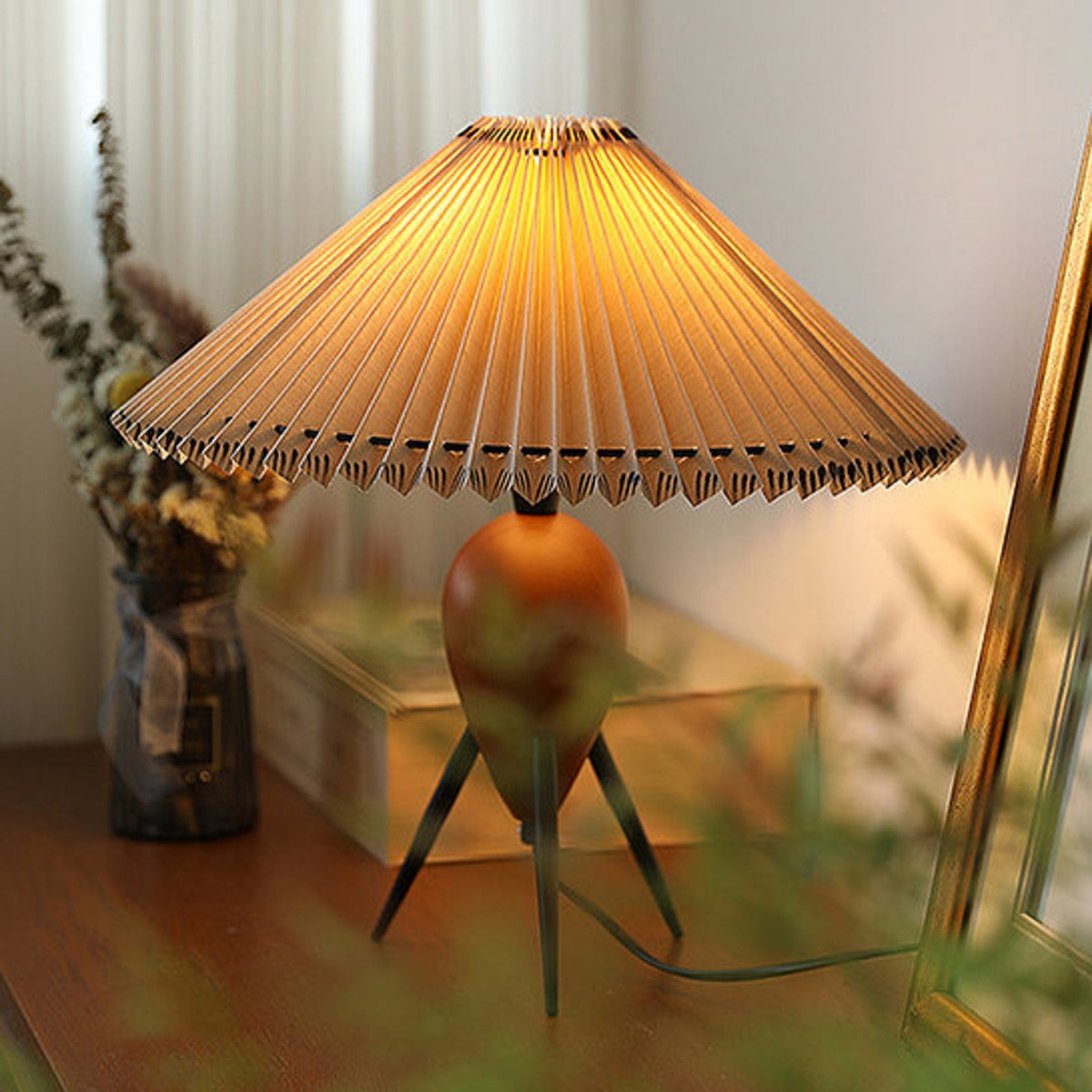Soft-focus close shot of a lit Clérachic table lamp on a wood shelf, flowers and a gold frame blurred behind it.