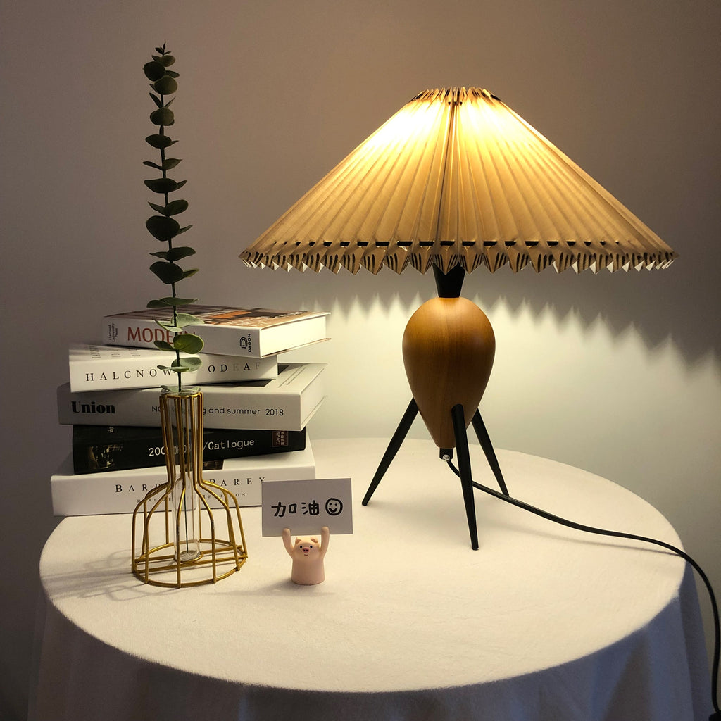 Glowing amber, a Clérachic table lamp on a draped round table with books and a gold wire vase, in a dim room.
