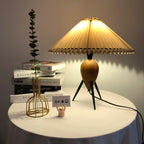 Glowing amber, a Clérachic table lamp on a draped round table with books and a gold wire vase, in a dim room.