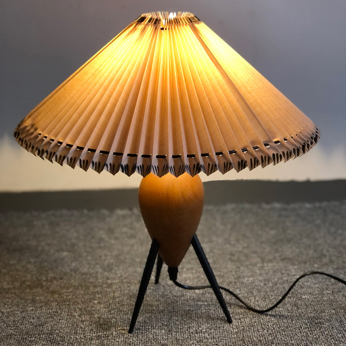 This Clérachic table lamp lit on grey carpet, light shining through the pleats above its wood base and slim black legs.