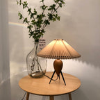 Beside a glass demijohn of leafy branches, a lit Clérachic table lamp stands on a pale round wooden table.