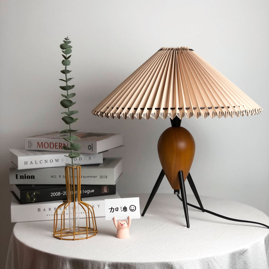 Seen at table height, a Clérachic table lamp stands by a gold wire vase and stacked books, its shade glowing faintly.