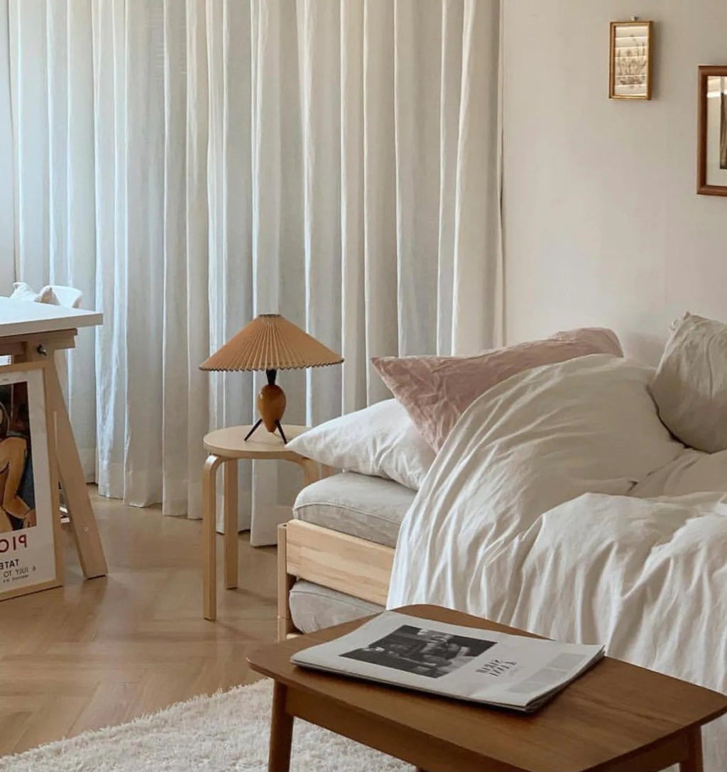 Seen across a bedroom, a Clérachic table lamp stands on a small stool by a bed with white linen and soft cushions.