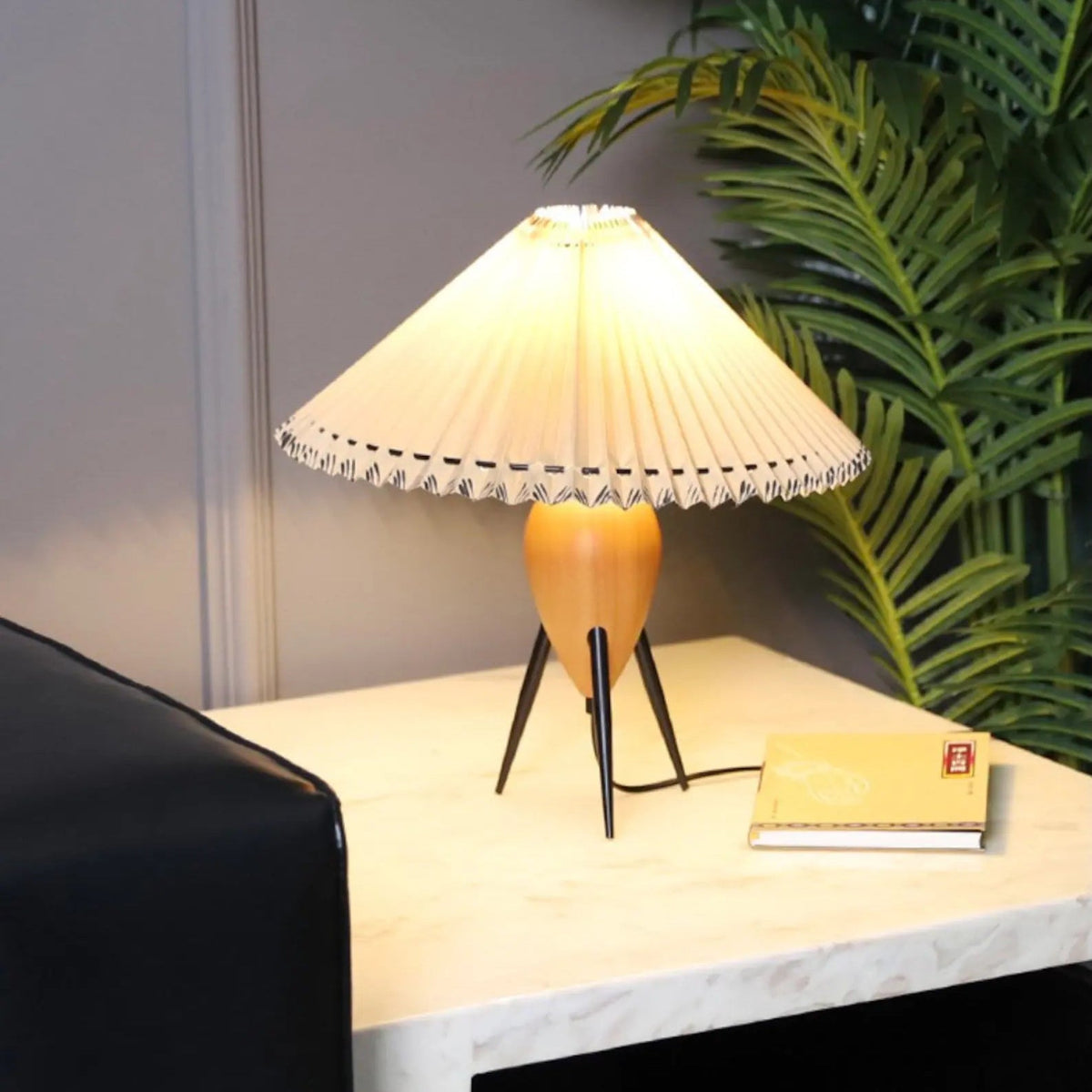 A lit Clérachic table lamp on a white marble-topped side table, a leafy palm behind it and a yellow notebook nearby.