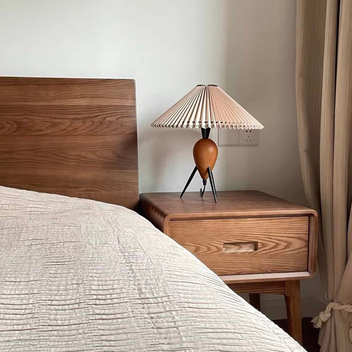 This Clérachic table lamp stands on a pale oak bedside table next to a wooden headboard and linen curtains.