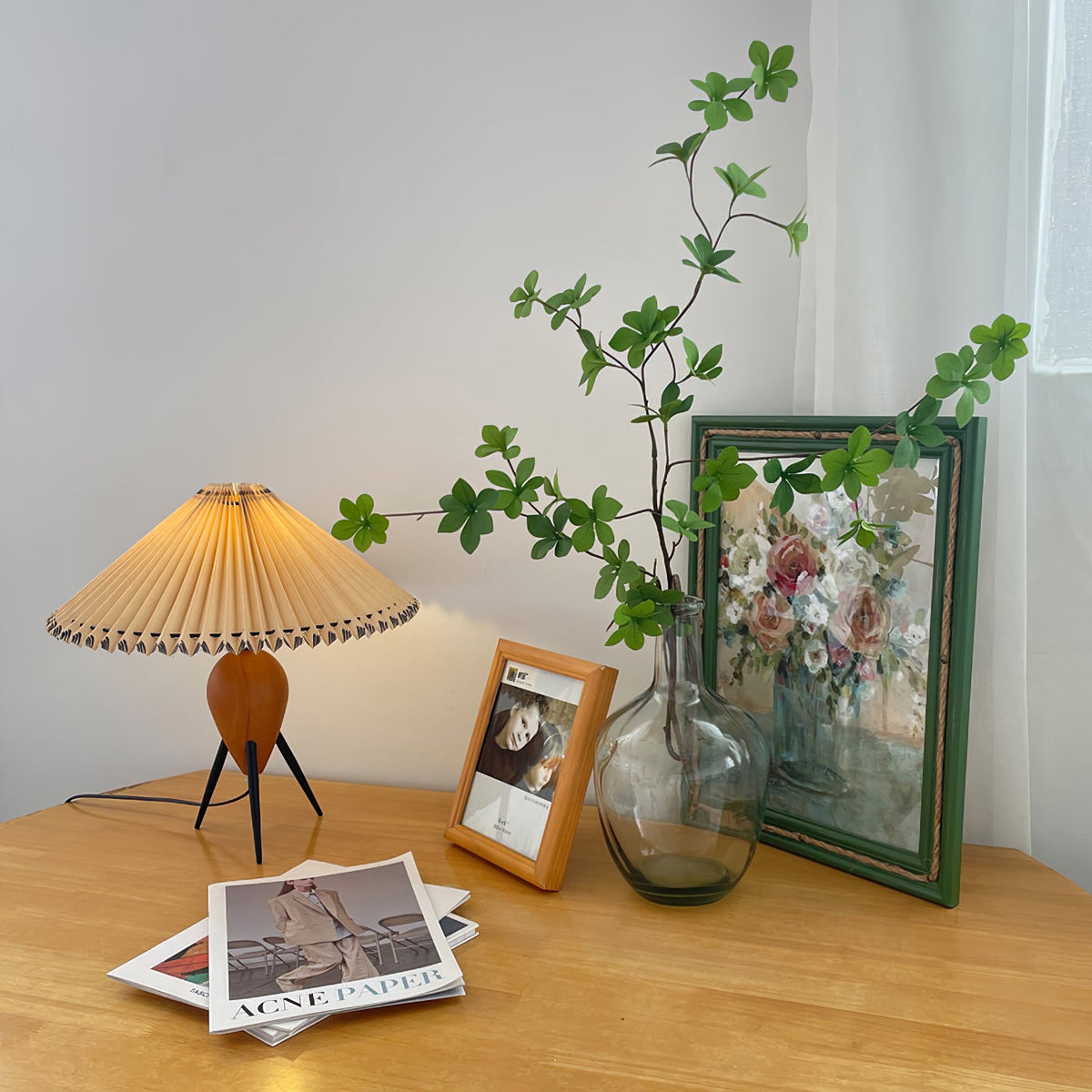 A Clérachic table lamp on a honey-coloured wood table, with a framed photo, a glass vase of green sprigs and magazines.